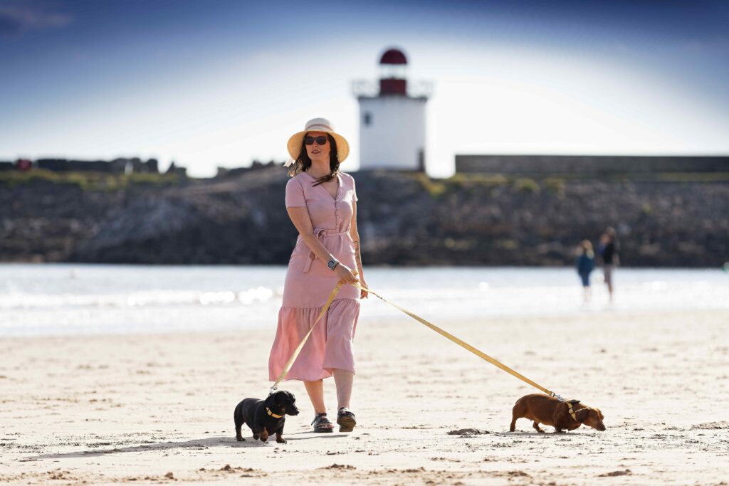 A lady wearing a pink dress and walking two dogs on a beach in Burry Port, Wales