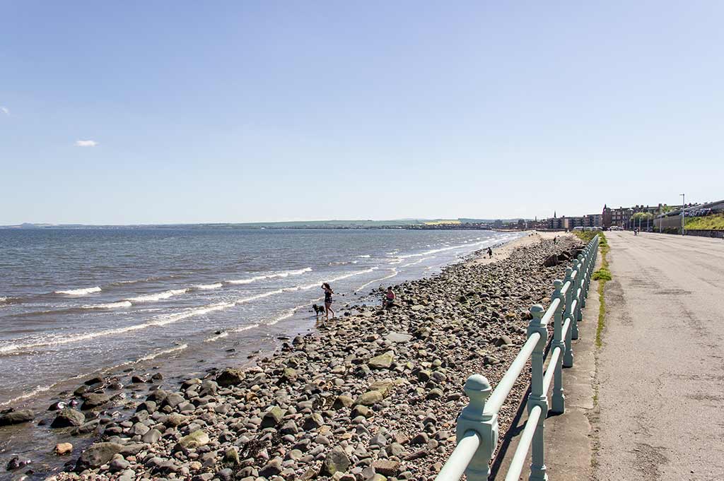 A wide shot of Portobello Beach; women play with two dogs on the pebbles by the shore