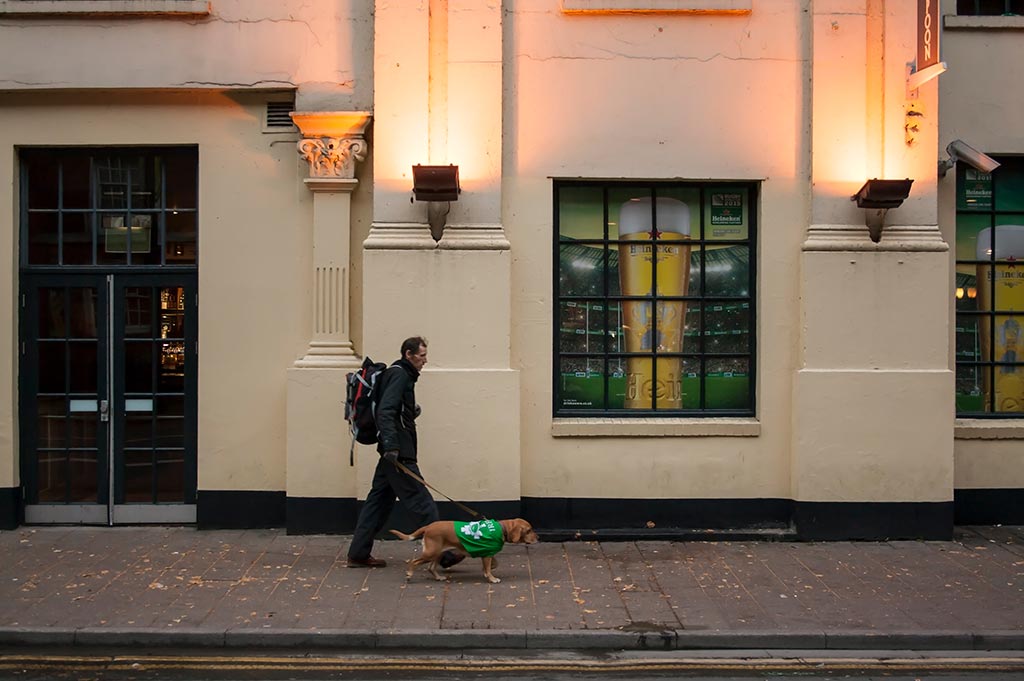 A man in a black coat walks a small brown dog in a green coat on a street in Cardiff