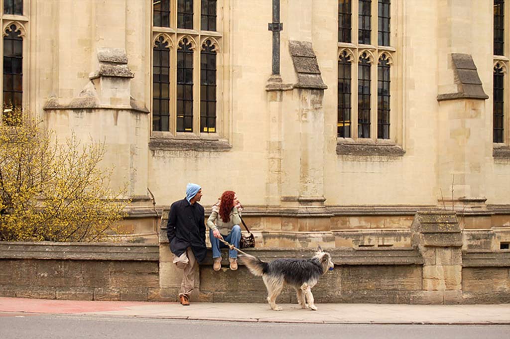 A man leans against a sandstone wall next to a woman who is seated on the wall, holding a large dog on a leash_ a grand sandstone building is in the background