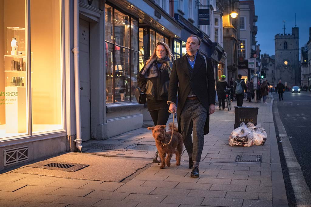 A man walks a brown dog down a pavement on High Street, Oxford, with a woman a few paces behind