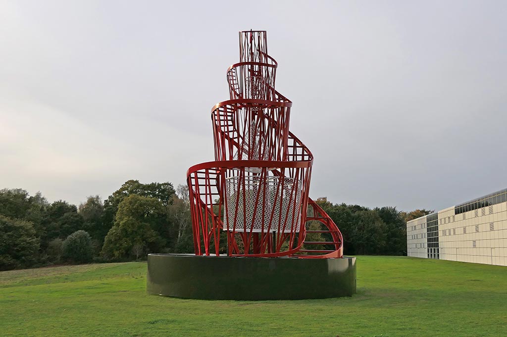 A statue comprised of red metal spirals stands in the centre of a green field in Sainsbury Centre