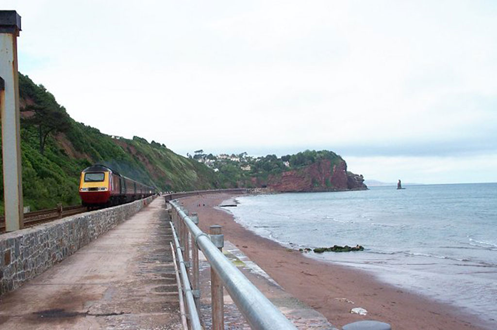 A train passes by a large body of water with rolling green hills in the background on Exeter's Great Scenic Railway