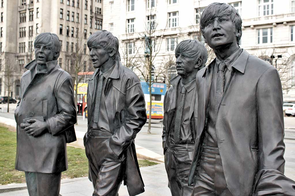 A waist-up shot of a bronze statue of The Beatles, with Liverpool's cream buildings in the background