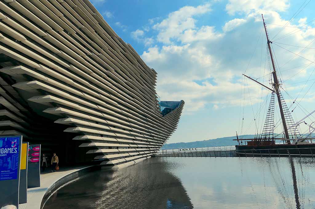 A wide shot of the VA in Dundee, a modern, architectural building set on the water, with an old sailing boat floating the background