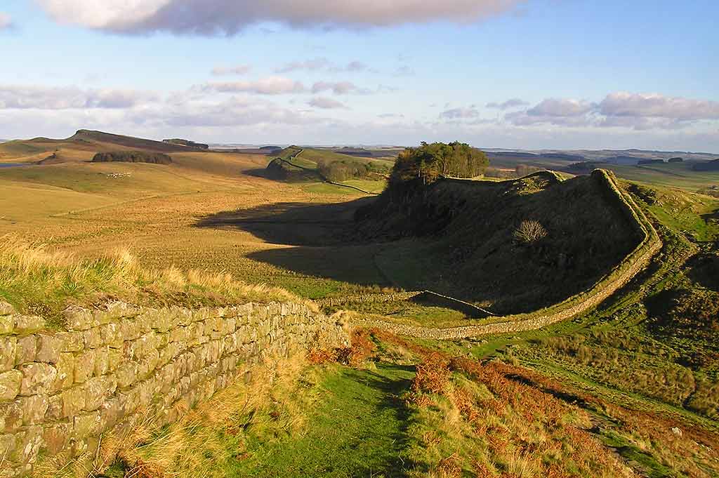 A wide view of Hadrians Wall at golden hour, showing the ancient stone walls cutting through lush green fields