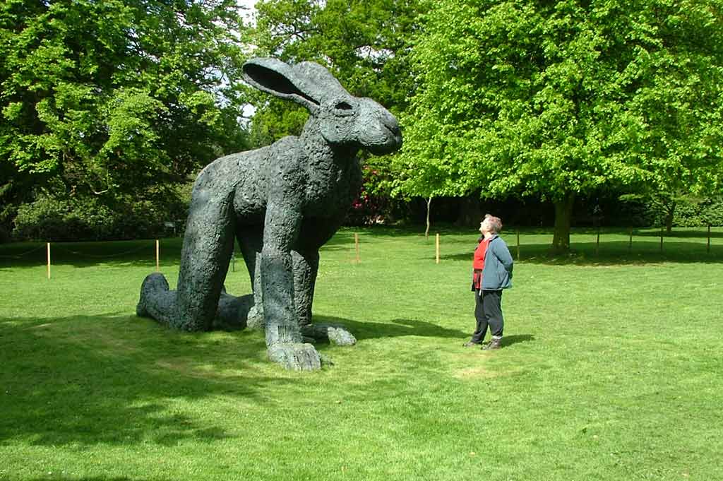 A woman stands next to a large sculpture depicting a hybrid rabbit-human form in the middle of Yorkshire Sculpture Park