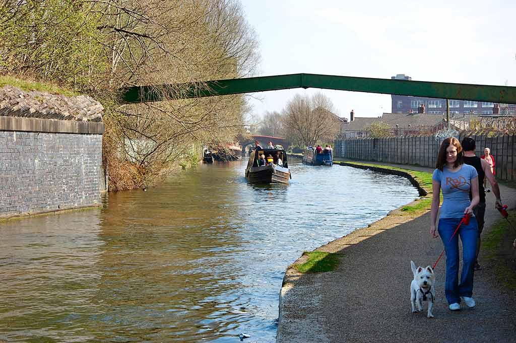 A woman walks a white dog along a pathway by a canal in Leeds_ a boat floats on the water