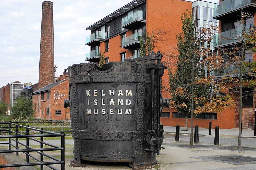 An antique steel ladle with the words _Kelham Island Museum_ affixed to it, in front of converted industrial buildings