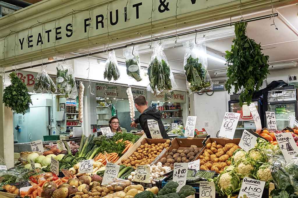 An indoor market stall selling fruit and vegetables at Cardiff Central Market with a man and a woman speaking in the background