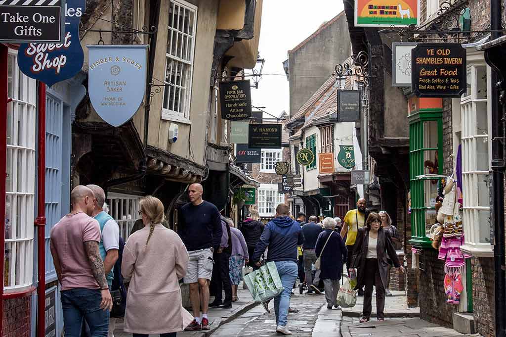 Crowds walk along The Shambles, a busy shopping street in York paved in cobblestones with quaint buildings either side