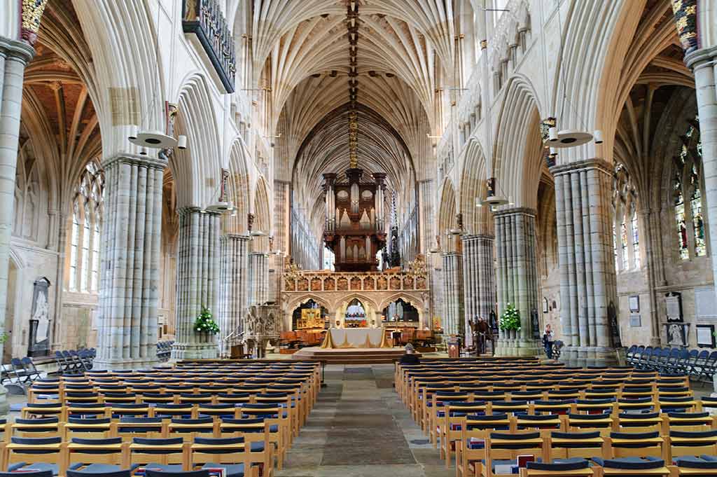The interior of Exeter Cathedral, with thick stone columns, impressive arched ceilings and rows of wooden seats lead to an ornate alter