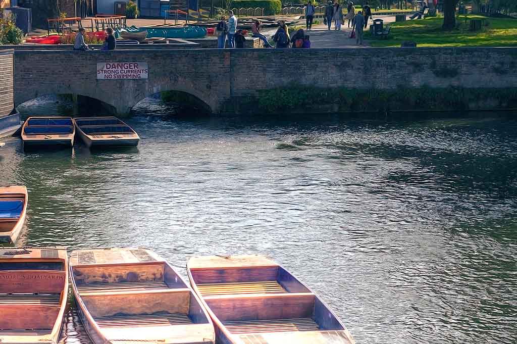 Wooden boats float on the River Cam in the foreground, while people cross a stone bridge in the background